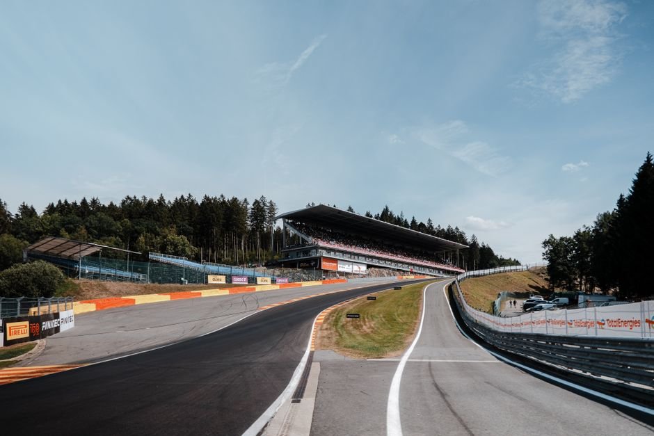 A dynamic view of a race track with a grandstand under a clear blue sky, surrounded by trees.